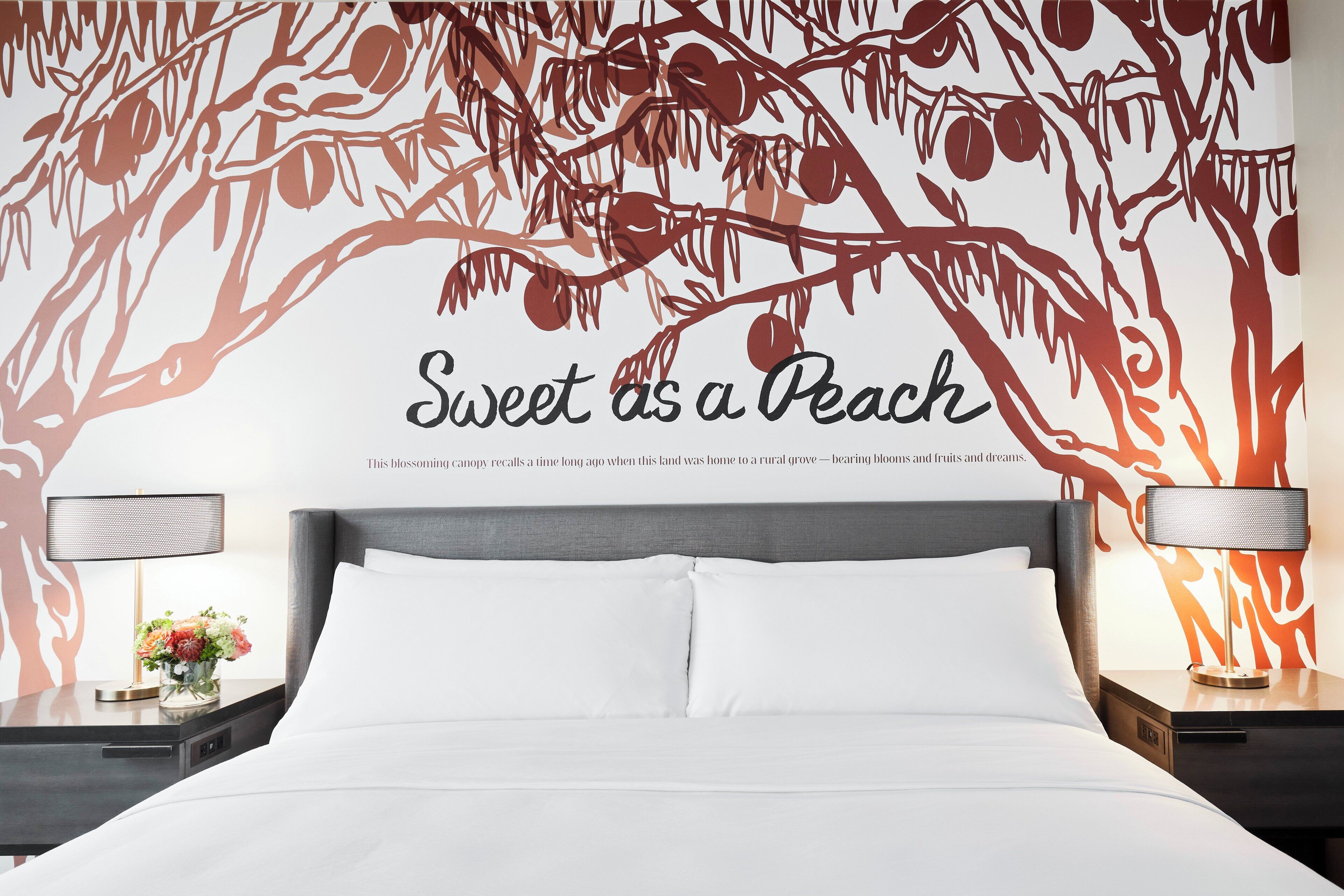 A bedroom with a large bed, two bedside tables, and a wall with a tree branch design and the phrase 'Sweet as a Peach.'