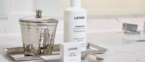 A bathroom with a Lather moisturizer bottle, a gentle face soap bar, and a metal container on a marble countertop.