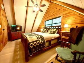 A cozy attic bedroom with a sloped ceiling, a bed with patterned bedding, a chair, a desk with a lamp, and a television on a dresser. - ★Cozy Log Cabin★ Secluded, modern amenities, mins to dwtwn Blueridge. Pets OK! (Blue Ridge)