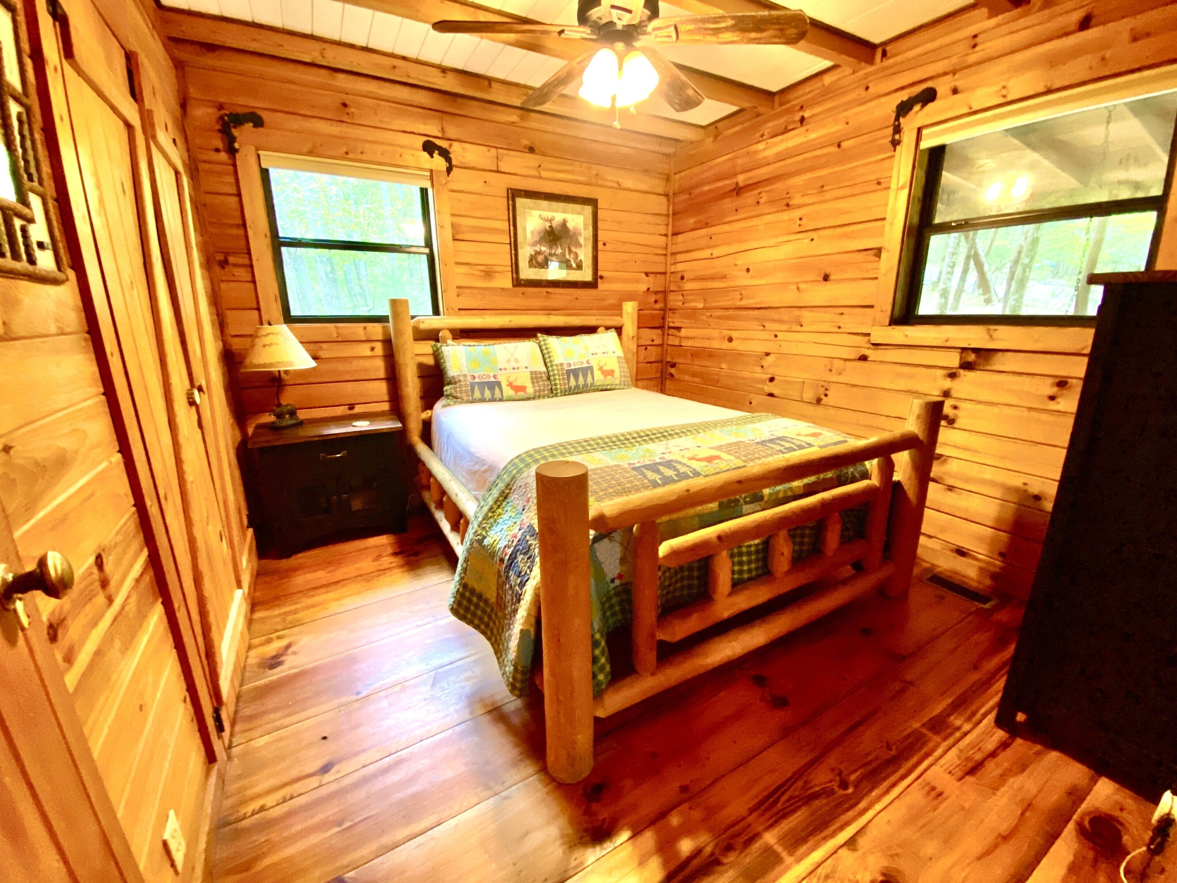 A wooden cabin bedroom with a bed, nightstands, a ceiling fan, and windows.