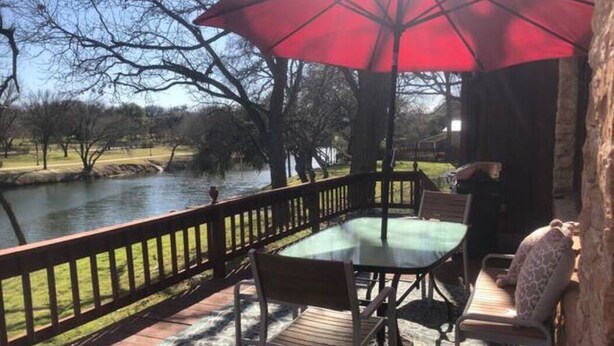 Una terraza con mesa y sillas de vidrio, un paraguas rojo y vista al río.