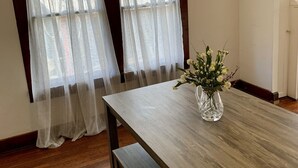 Un comedor con mesa de madera y banco, dos ventanas con cortinas translúcidas y un candelabro.