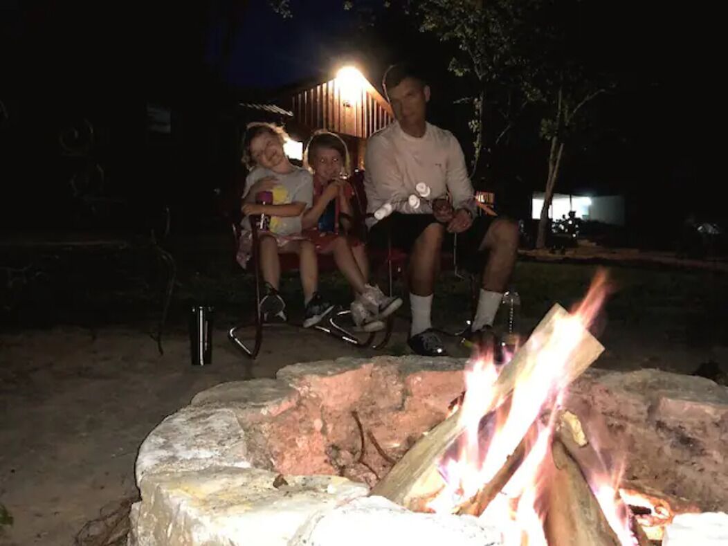 Ein abendliches Außenszenario mit einem Feuerkorb, einer Person, die auf einem Stuhl sitzt, und einer stehenden Person.