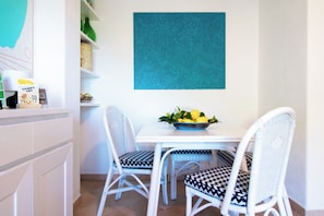 Un coin repas avec une table blanche et deux chaises blanches dotées de coussins à motifs, une décoration murale bleue texturée au-dessus, et un bol de citrons posé sur la table.