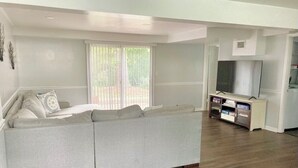 A modern living room with a grey sectional sofa, a TV stand with a flat-screen TV, and a large window with blinds. - Sleeps 10! Brand new kitchen & main bathroom! Highly sought after 4bd,3ba. (Smyrna)