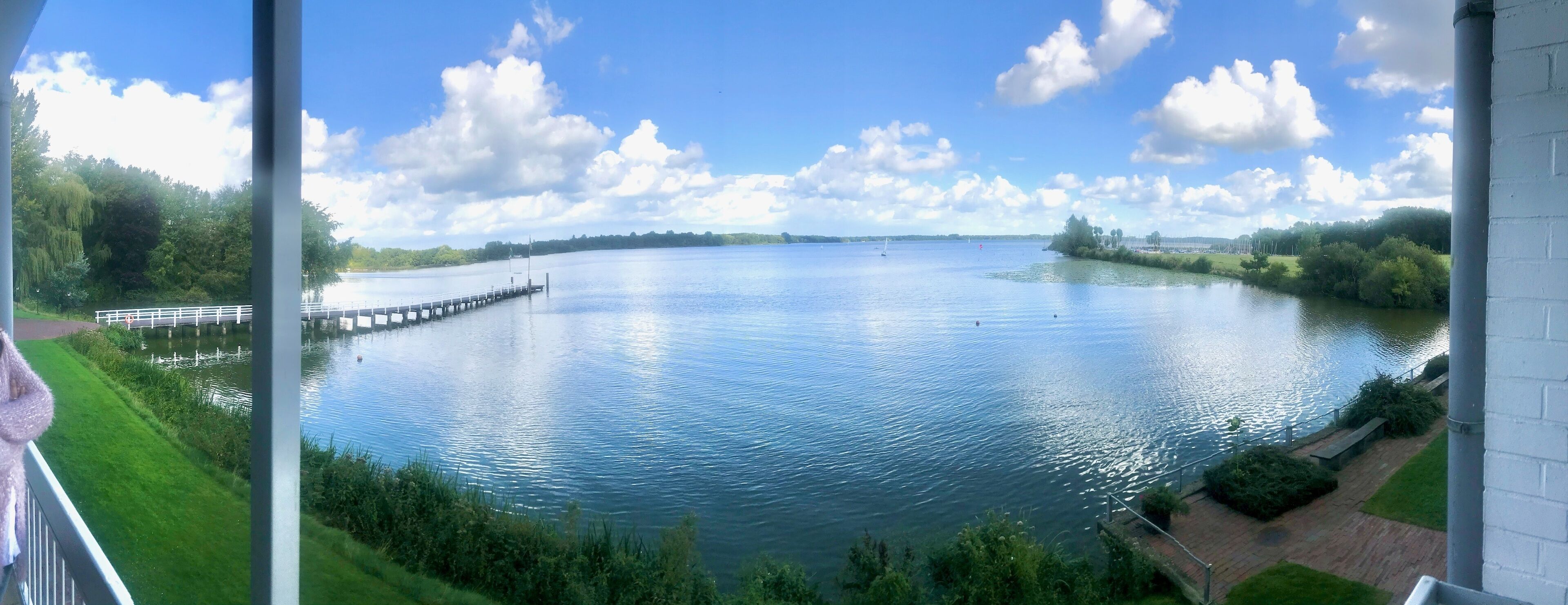A view of a calm lake with a dock extending into the water, surrounded by greenery and a clear sky.