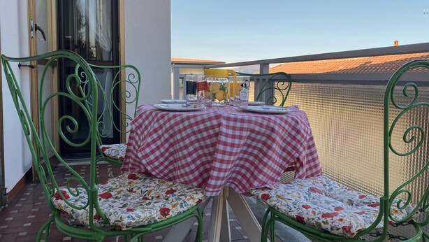 Un balcon extérieur avec une table dressée pour deux personnes, agrémenté d’une nappe à carreaux et de coussins à motifs floraux.