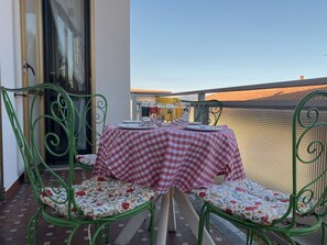 Un balcon extérieur avec une table dressée pour deux personnes, agrémenté d’une nappe à carreaux et de coussins à motifs floraux.