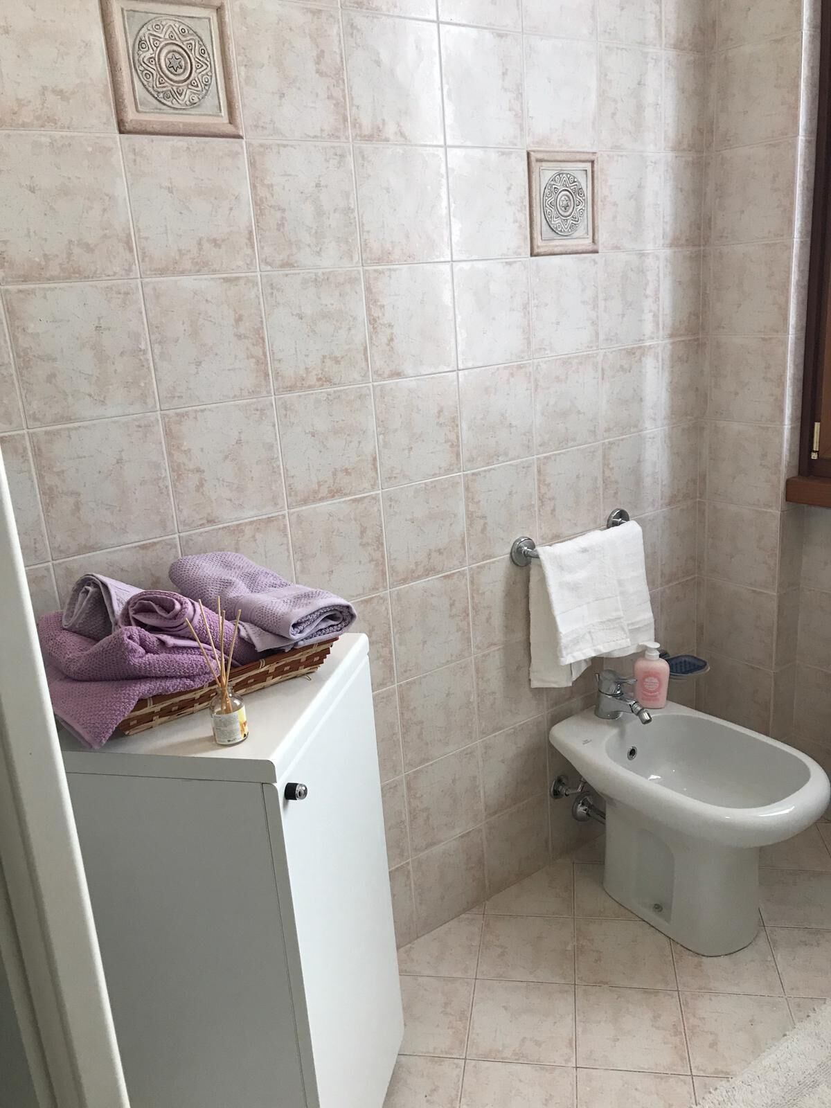 Une salle de bain carrelée de carreaux beige, avec des toilettes blanches, un bidet et un meuble contenant des serviettes pliées et un diffuseur de parfum à bâtonnets.