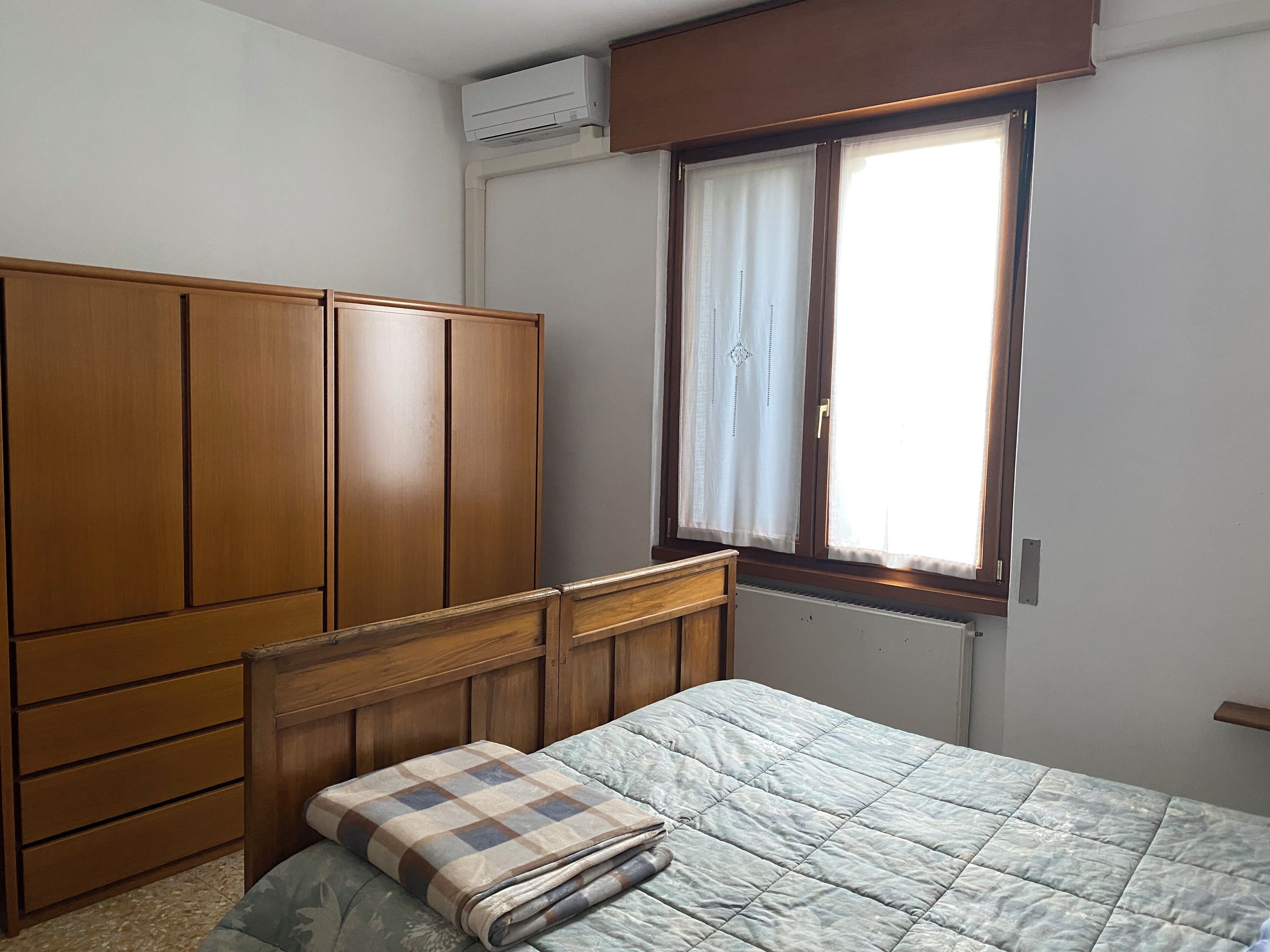 A bedroom with a wooden wardrobe, a bed with a checkered blanket, and a window with curtains.