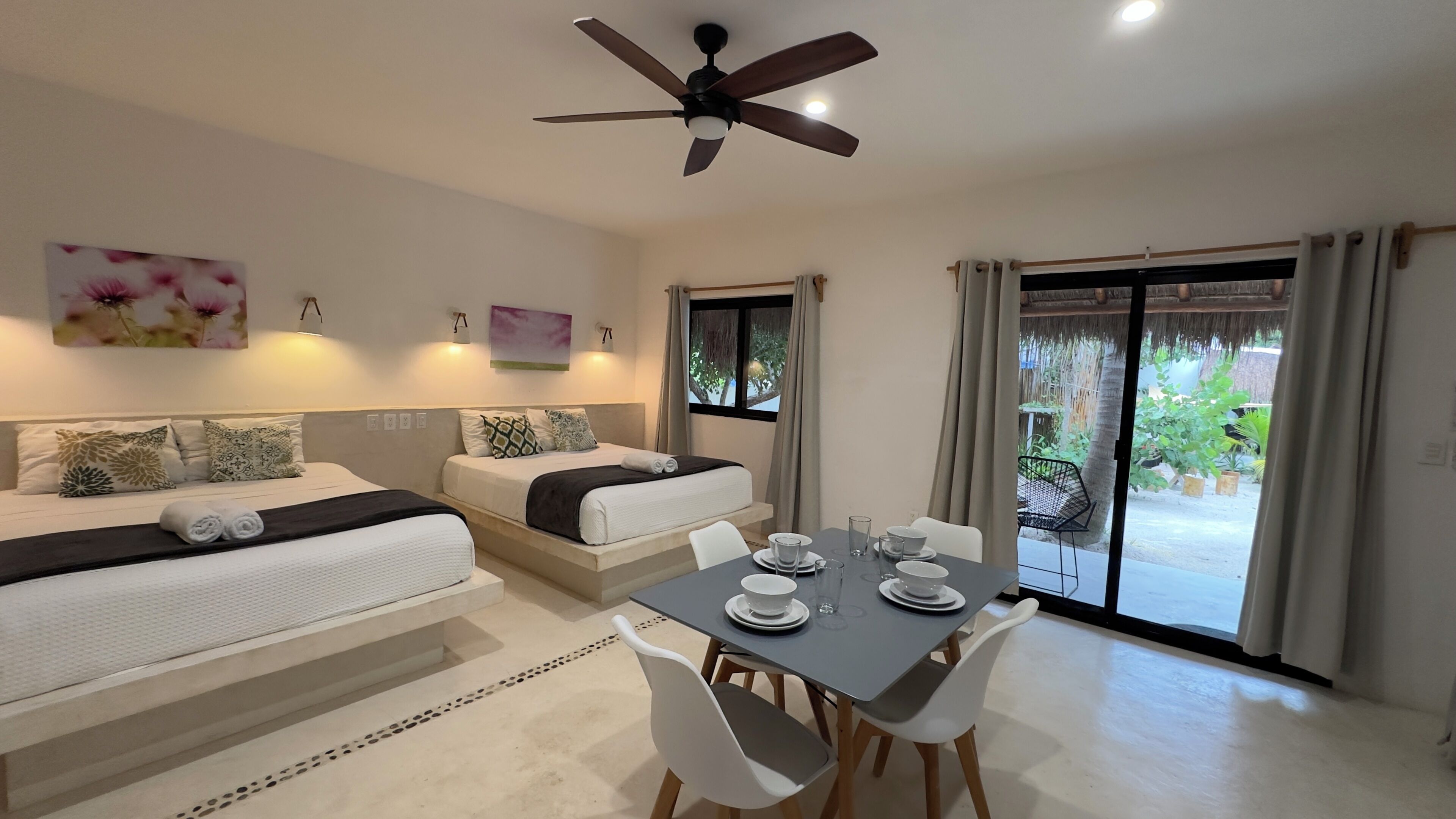 A hotel room with two beds, a dining table with chairs, a ceiling fan, and a view of the outdoors through a sliding door.