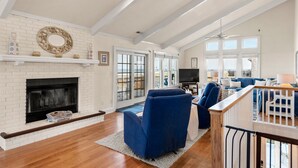 A spacious living room with a white brick fireplace, blue seating, and large windows offering a view. - Chez la Mer – Oceanfront 5BR Sanderling Home with Private Walkway & Big Views (Duck)