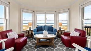 A room with a large bay window, a red leather sofa, a blue sofa, and a round coffee table with a decorative vase. - Chez la Mer – Oceanfront 5BR Sanderling Home with Private Walkway & Big Views (Duck)