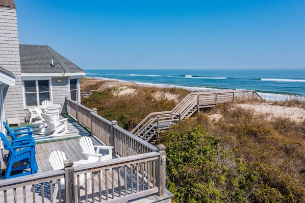 A wooden deck with white Adirondack chairs overlooking a beach.