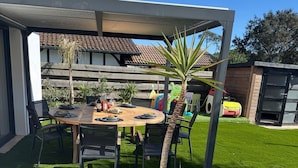 En overdækket terrasse med et træbord og stole, en lille palme og en rød legetøjsbil i baggrunden.
