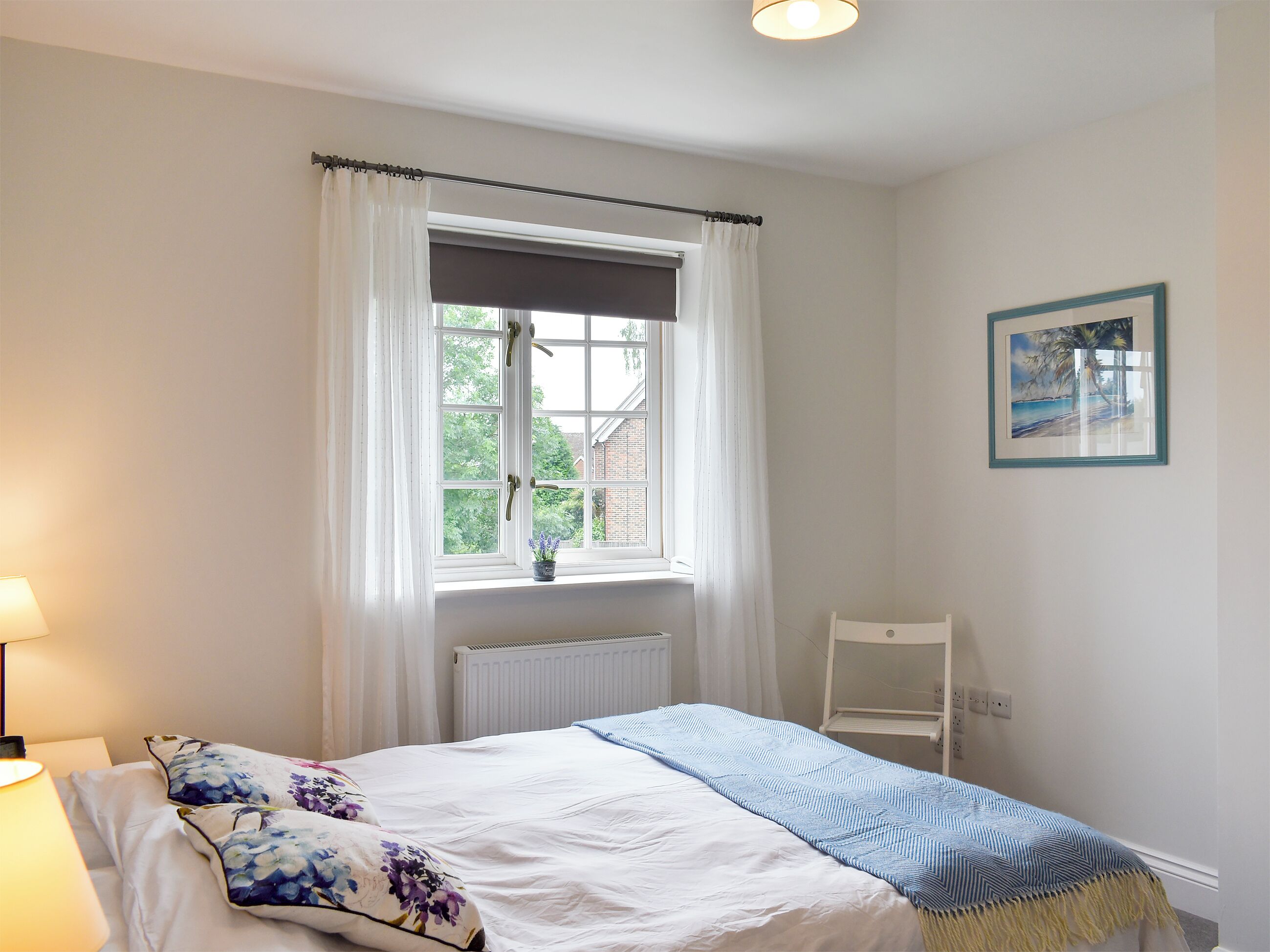 Un dormitorio con cama, ventana con cortinas, radiador, una silla e imagen colgada en la pared.