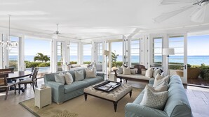 A spacious living room with a large window overlooking the ocean, a ceiling fan, and a dining area with a view. - Santosha Villa Estate, Private Staffed Beach Resort All to Yourself, Sleeps 21 (Long Bay Village)
