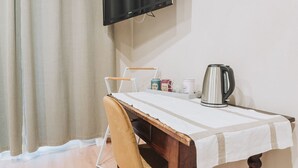 A small room with a wooden table, a chair, a television mounted on the wall, and a kettle on the table.