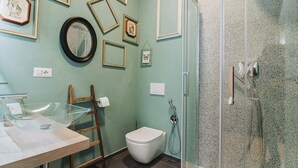 A bathroom with a glass shower enclosure, a toilet, a sink with a wooden frame, and various framed pictures on the wall.