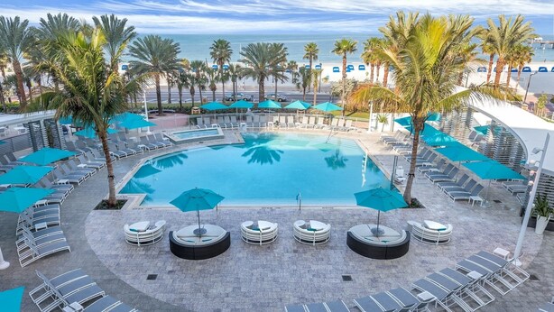 A pool area with lounge chairs and umbrellas, surrounded by palm trees and a view of the ocean.