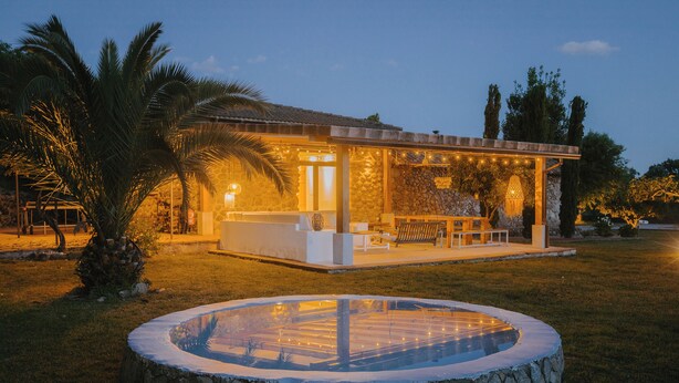 A modern outdoor pool area with a stone fountain, surrounded by a covered seating area and palm trees.
