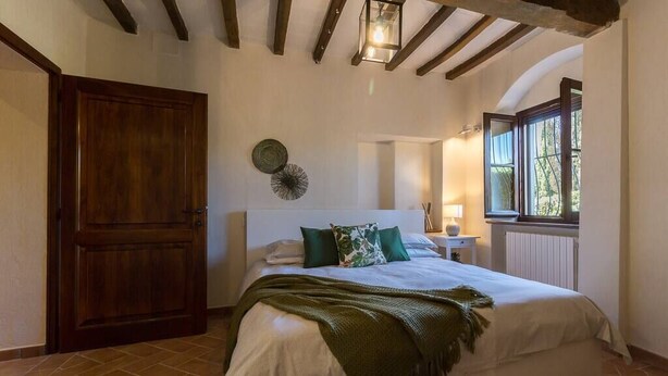 Une chambre avec un grand lit, une porte en bois, une petite table avec une lampe et une fenêtre donnant sur un paysage verdoyant.