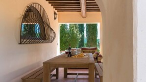 Une table en bois avec un banc, un élément décoratif en métal fixé au mur, et une vue sur un jardin avec un canapé et des coussins.