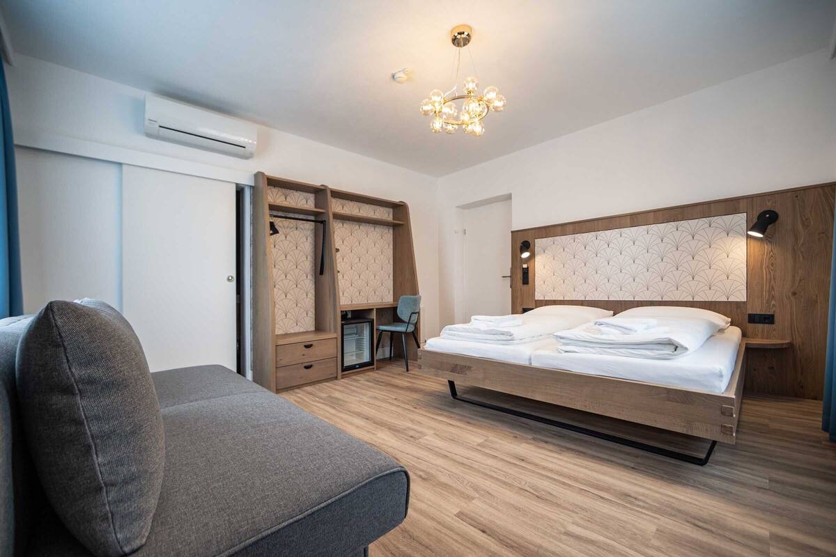 A modern hotel room with a wooden bed, a grey sofa, a desk, and a wardrobe.