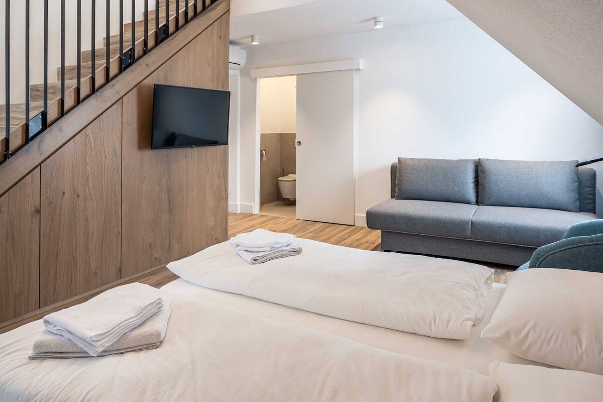 A modern living room with a grey sofa, a flat-screen TV mounted on a wooden wall, and a staircase leading to an upper level.