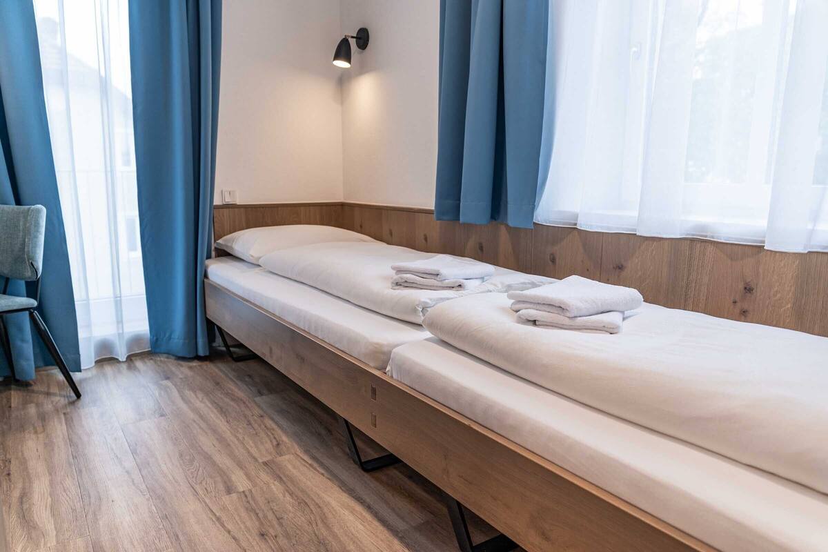 A single bed with white linens and a wooden headboard in a room with a wooden floor and blue curtains.
