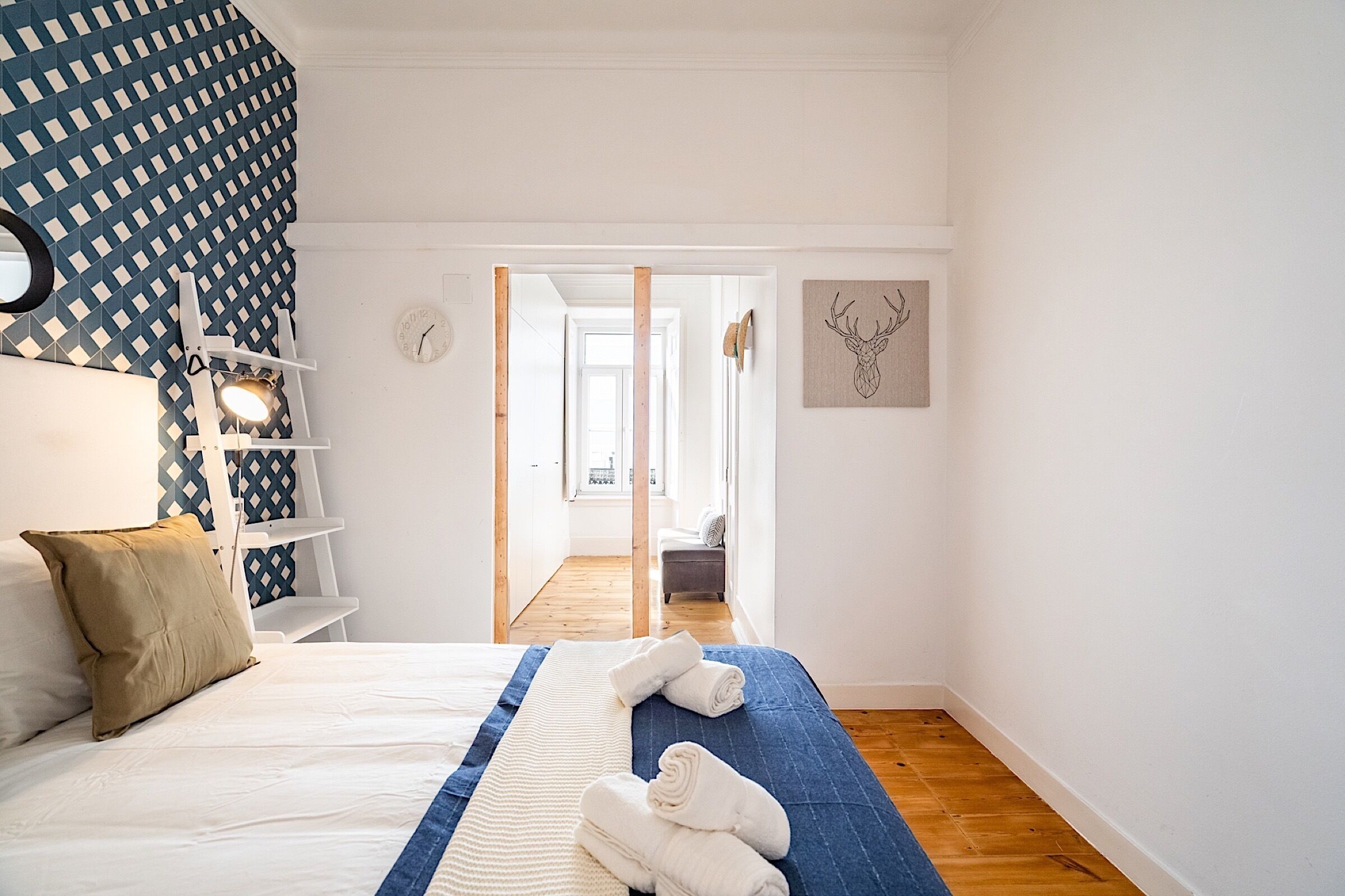 A bedroom with a bed, a blue and white striped blanket, a wooden floor, a white wall with a clock, a shelf with decorative items, and a door leading to another room.