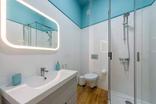 Une salle de bain moderne avec une douche à parois de verre, un grand miroir et un lavabo blanc doté d’un distributeur de savon bleu.