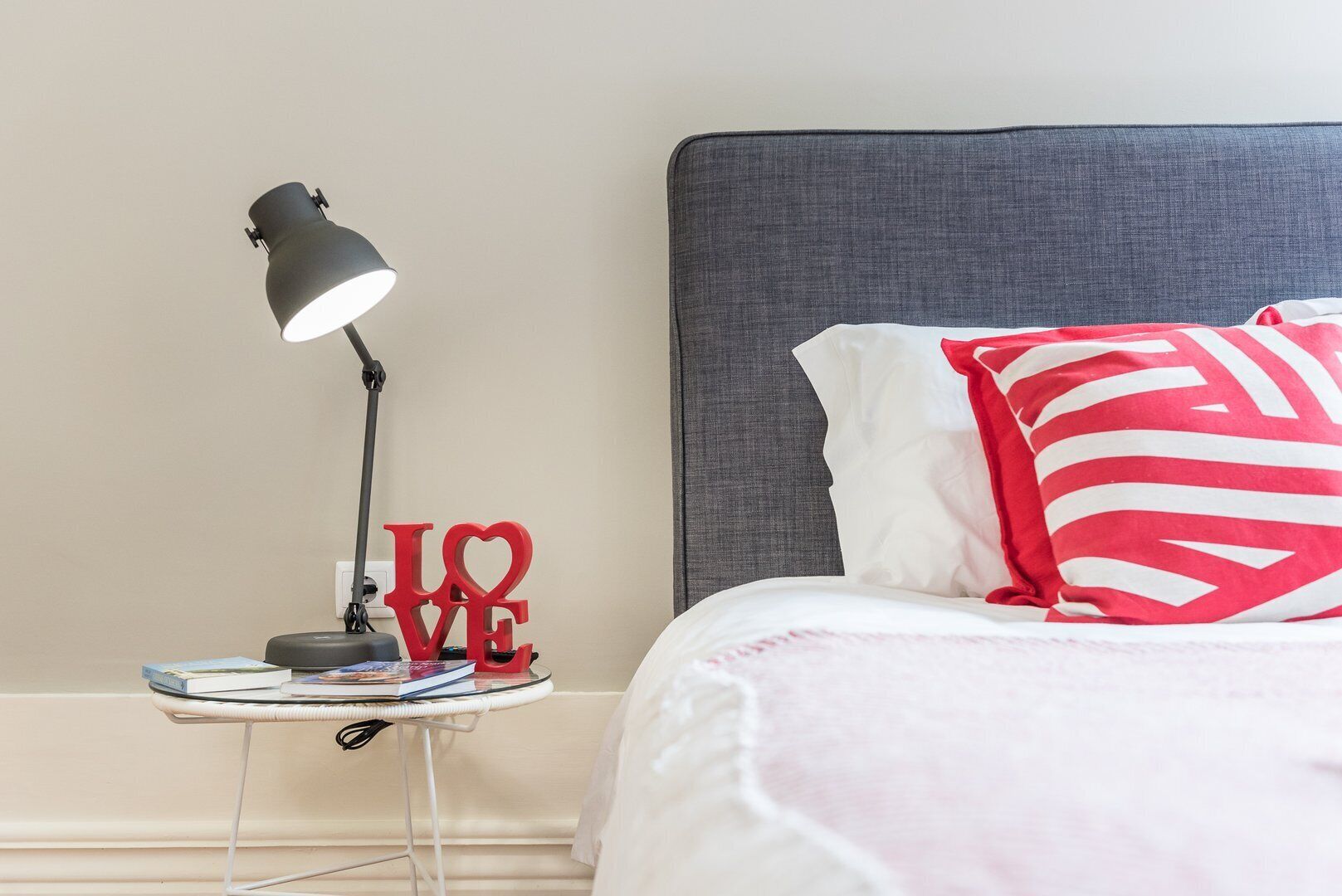 Un lit bien fait, avec une tête de lit grise, une lampe de chevet et une petite table sur laquelle sont posés des livres et un objet décoratif rouge représentant le mot « LOVE ».