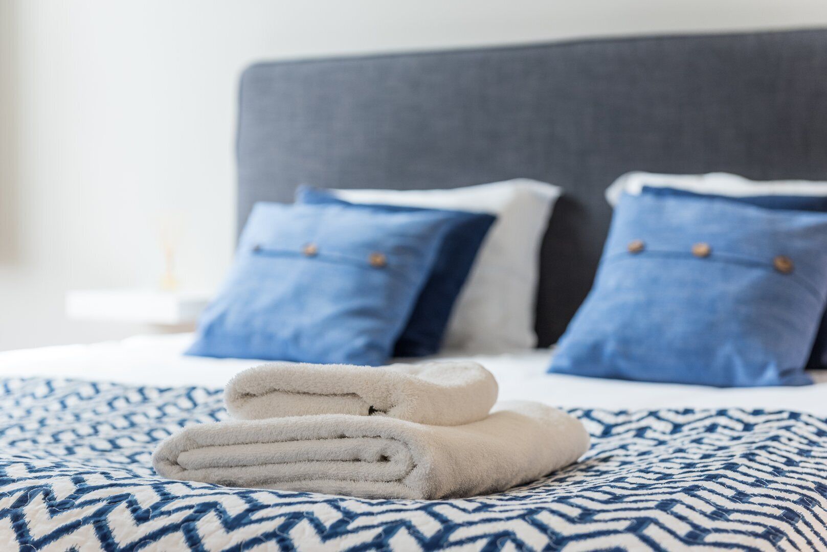 Una cama bien hecha con ropa de cama de diseño azul y blanco, dos almohadas azules con botones de madera, y una toalla blanca doblada sobre la cama.