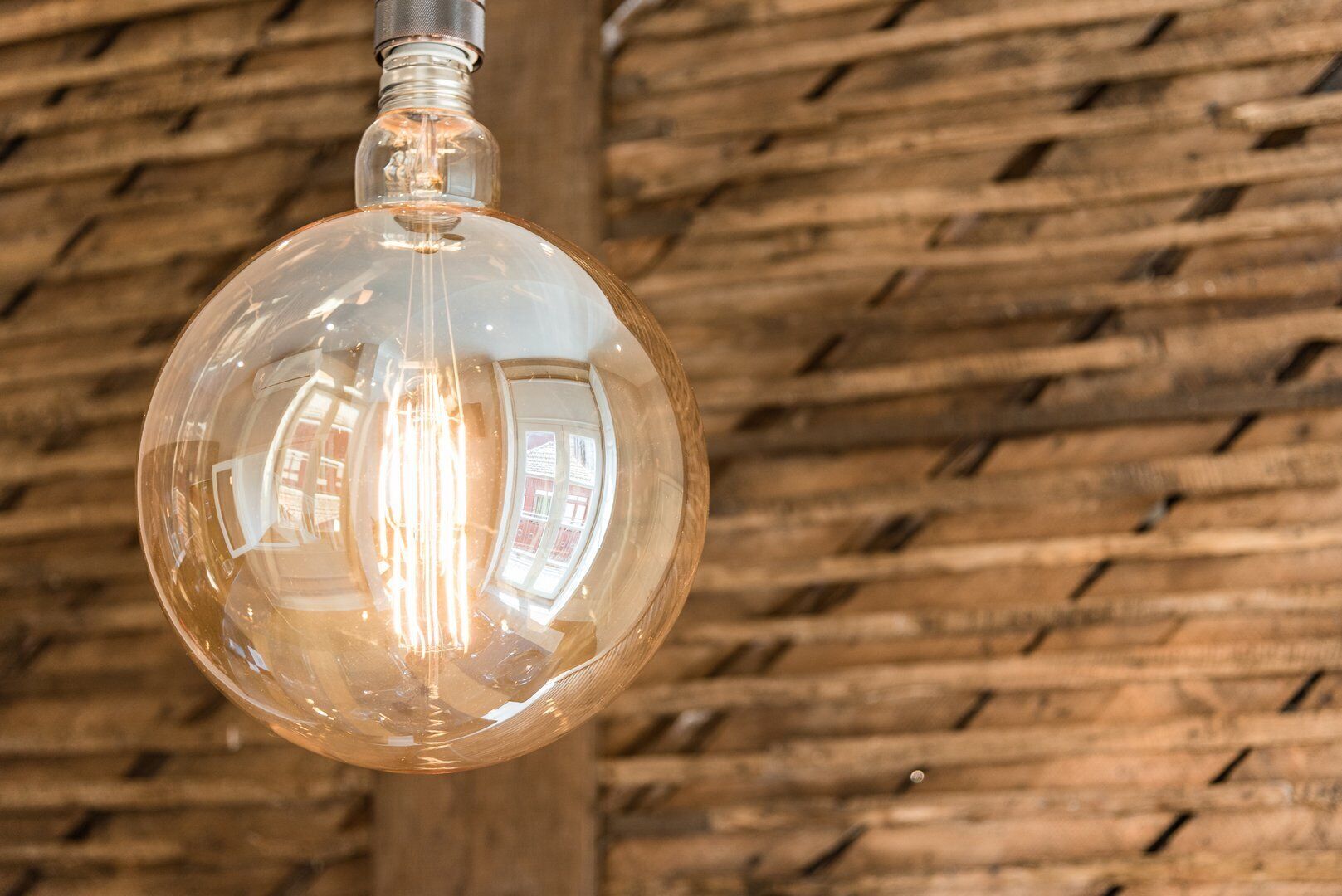 A clear glass pendant light with a metallic base, hanging against a textured wooden wall.