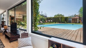 A modern living room with a large window overlooking a swimming pool, wooden deck, and outdoor furniture. - GuestReady - Sunset Pool House (Esposende)