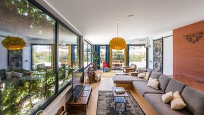 A modern living room with a large sofa, a coffee table, and a dining area with chairs. There is a large window with a view of greenery outside. - GuestReady - Sunset Pool House (Esposende)