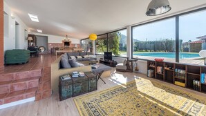 A spacious living area with a large window overlooking a swimming pool, a fireplace, and a modern kitchen in the background. - GuestReady - Sunset Pool House (Esposende)