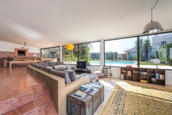 A spacious living area with a large window, a built-in fireplace, a pool, and a bookshelf. - GuestReady - Sunset Pool House (Esposende)