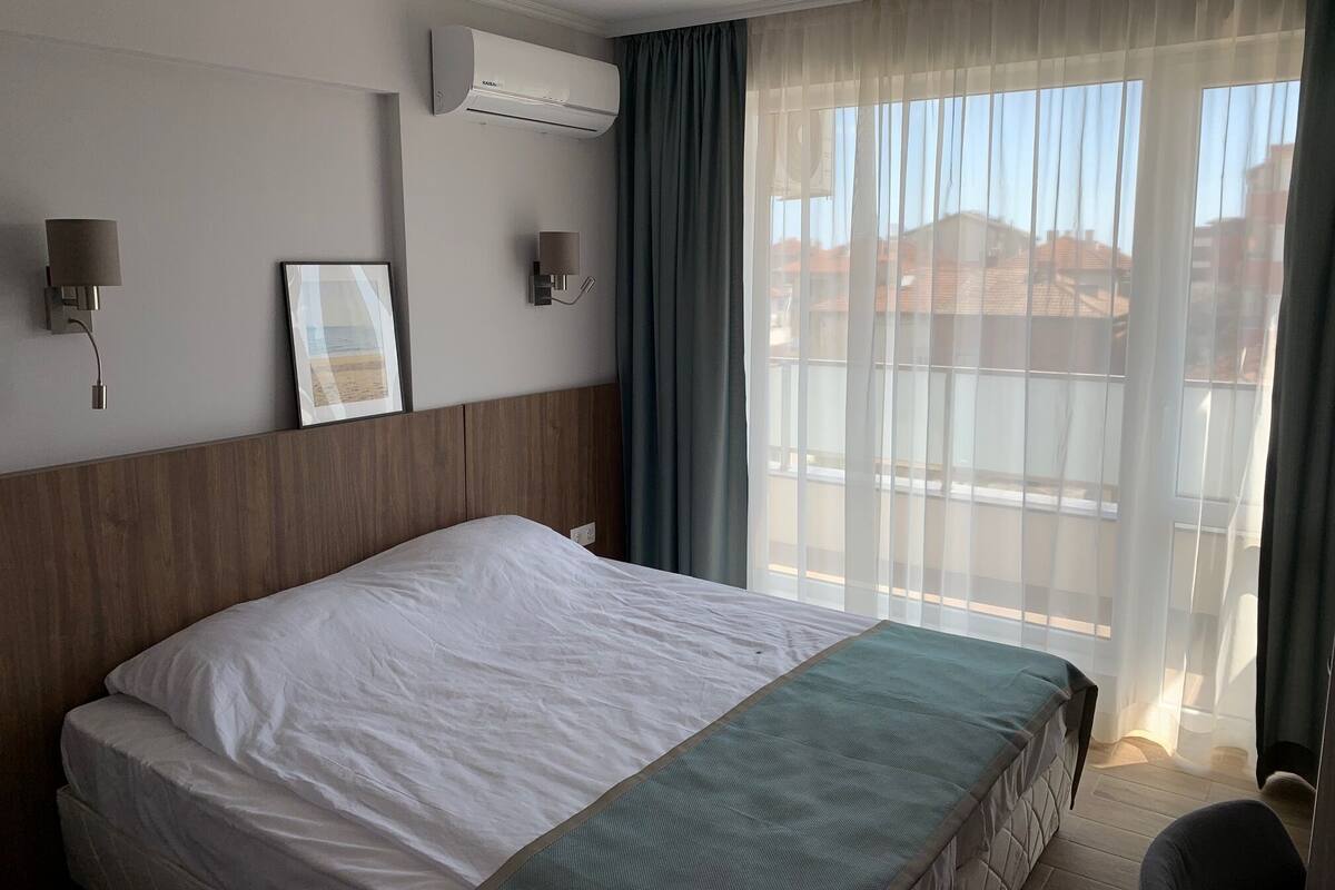 Una habitación de hotel con una cama grande, un equipo de aire acondicionado, una lámpara de pared y una ventana con cortinas translúcidas.