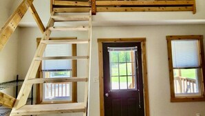Une chambre située dans un espace de type « loft », avec un escalier en bois, une porte noire et deux fenêtres équipées de stores.