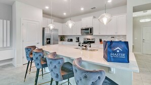 Uma cozinha moderna com armários brancos, uma ilha central com banquetas, eletrodomésticos de aço inoxidável e iluminação pendente.