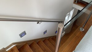 Una escalera de madera con barandilla de metal y balaustrada de vidrio, que conduce a una pared blanca con iluminación empotrada.