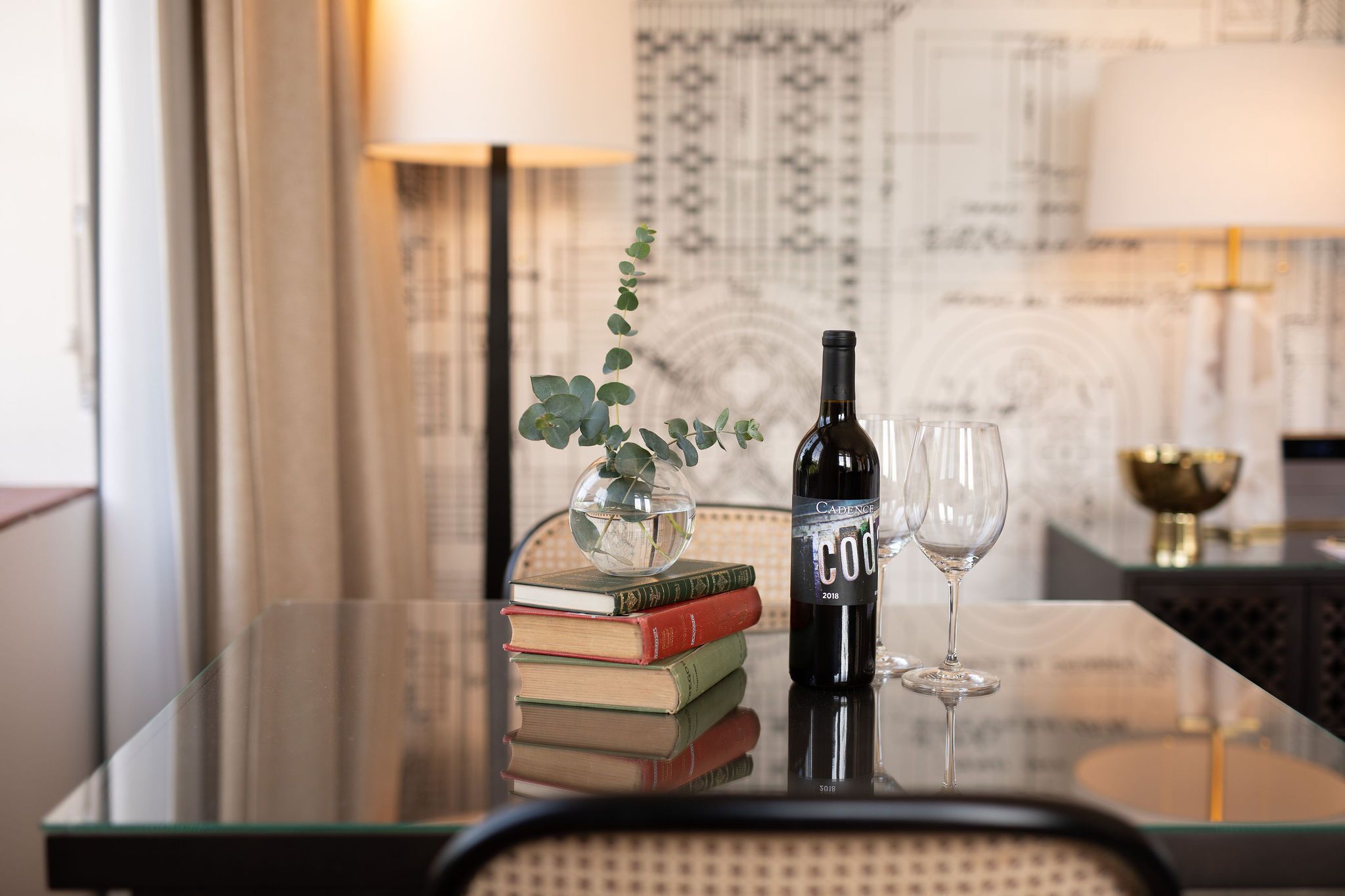 A glass table with a bottle of wine, a glass, and a vase with a plant. There are books stacked on the table.