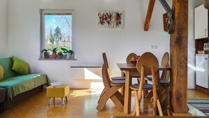 Un salon chaleureux avec un canapé vert, une table à manger en bois avec des chaises, un petit pouf jaune et une fenêtre agrémentée de plantes en pot.