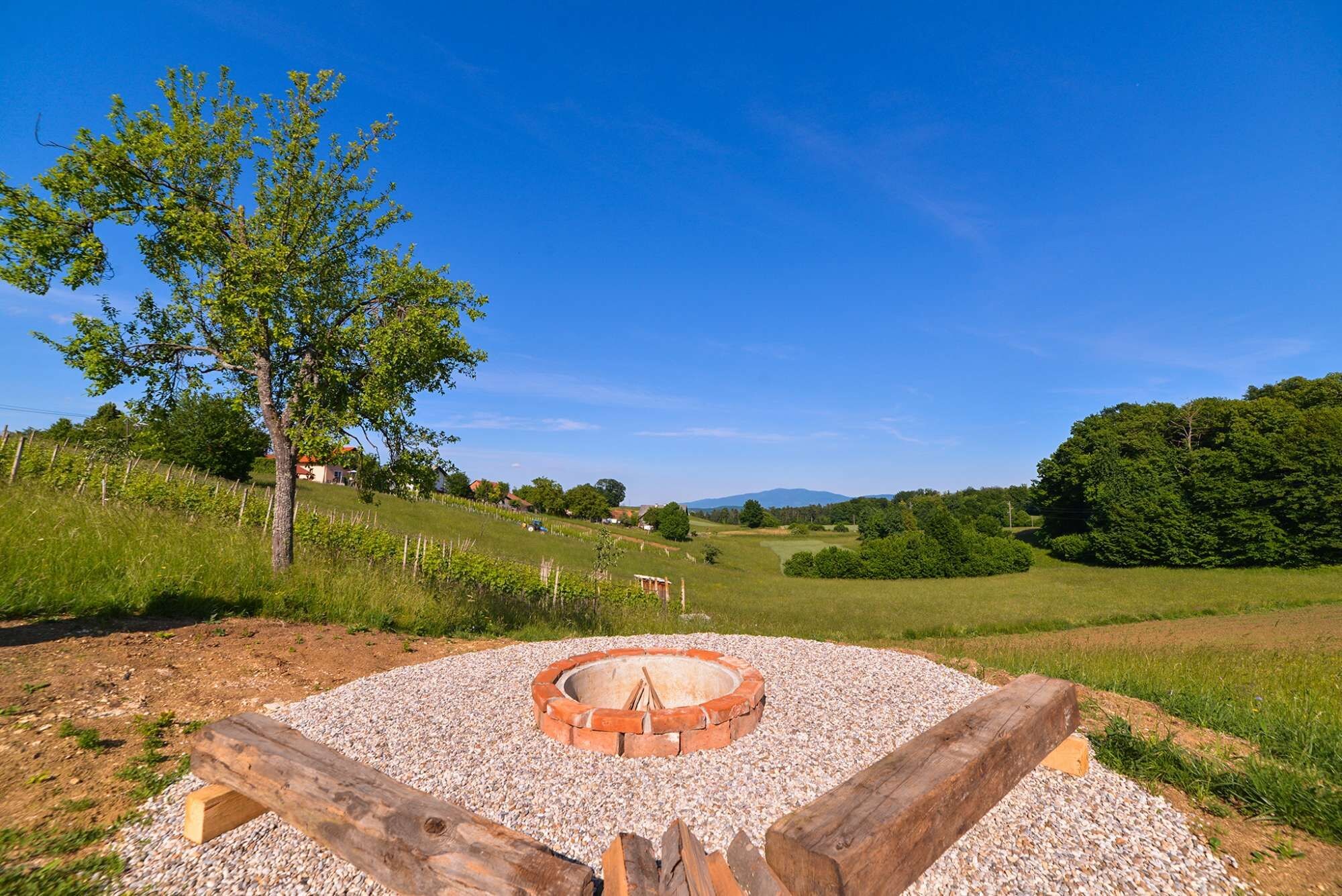 A circular fire pit with a gravel base, surrounded by a wooden fence, set in a grassy field with trees and houses in the background.