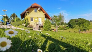 Une maison jaune avec un toit rouge, entourée d’herbe verte et d’arbres, sous un ciel bleu clair.