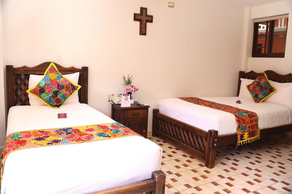 Habitación de hotel con dos camas, cada una con una colcha floral de colores y almohadas a juego. Cuenta con un cabecero de madera, una mesita de noche con un florero y un crucifijo colgado en la pared. El suelo es de baldosas y tiene una ventana con persianas.