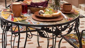 Una mesa redonda con un tablero de mosaico, rodeada de sillas, un jarrón con flores y un frutero.
