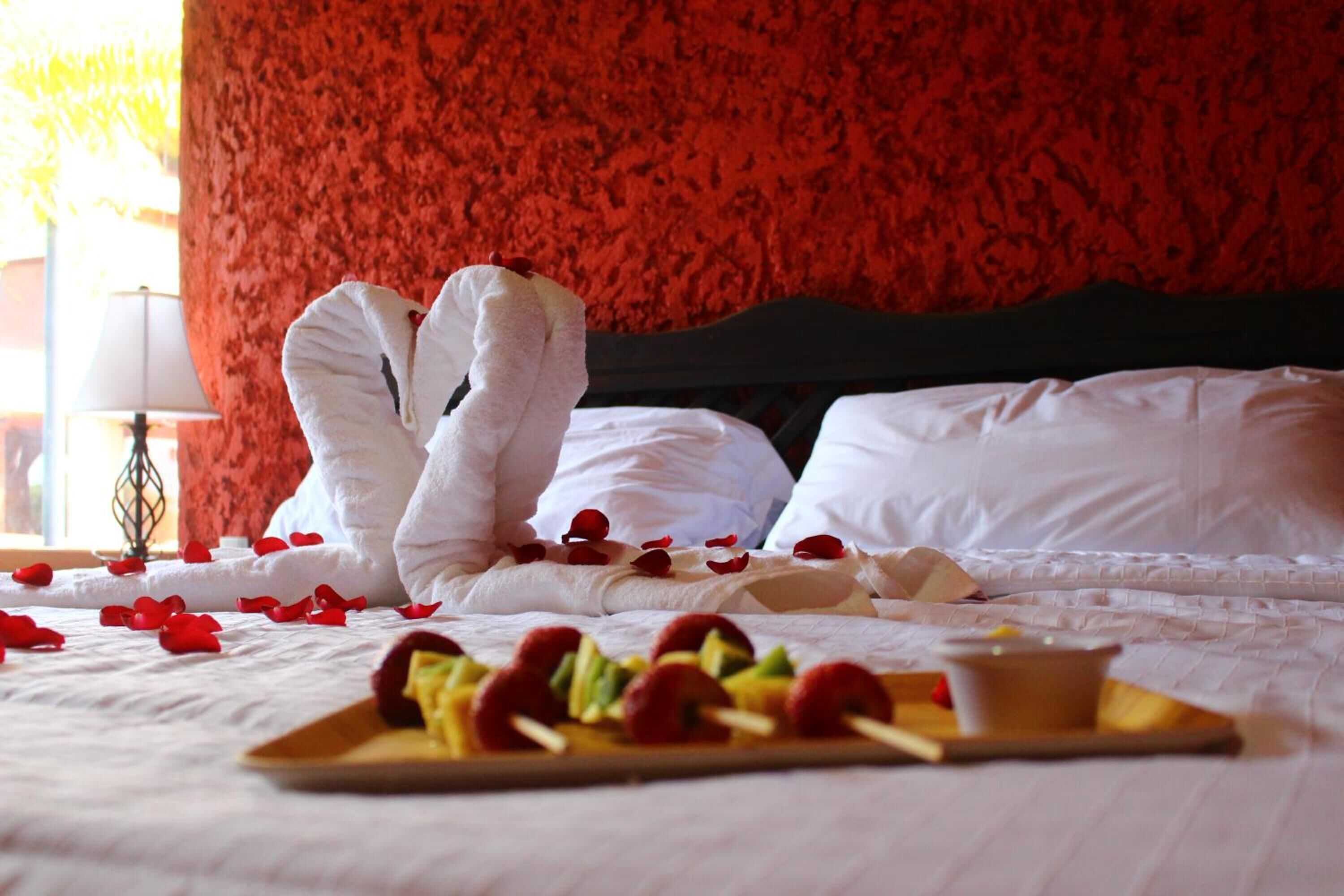 Un letto con cuscini bianchi e un vassoio con spiedini di frutta, un vassoio di legno e petali di rosa sparsi.